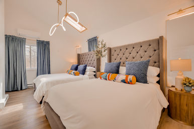 Bright, cozy bedroom interior with two tufted headboard beds in white linens, blue accent pillows and colorful bolster cushions, wooden nightstand with lamp, skylight and blue curtains.