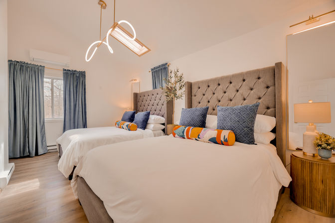 Bright, cozy bedroom interior with two tufted headboard beds in white linens, blue accent pillows and colorful bolster cushions, wooden nightstand with lamp, skylight and blue curtains.