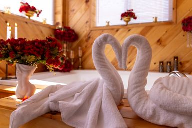 Romantic wood-paneled cabin bathroom with two white towel swans forming a heart on the tub edge, surrounded by red rose bouquets and lit candles.