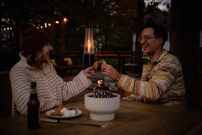 Two friends on a backyard deck at dusk, smiling and toasting s'mores over a tabletop fire pit with string lights and a patio heater in the background.