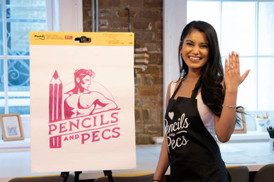 Smiling woman in a black apron shows her left hand with an engagement ring while standing beside an easel displaying a large pink illustration of a pencil and muscular torso logo in a bright art studio.