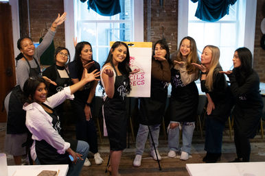 Smiling group of women in aprons posing around an easel, blowing kisses during a lively indoor painting workshop in a cozy brick-walled art studio.