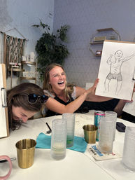 Two friends at an indoor sketching party laughing as one holds up a funny pencil drawing of a muscular man in underwear, with glasses, drink cans and art supplies on the table in a cozy studio setting.