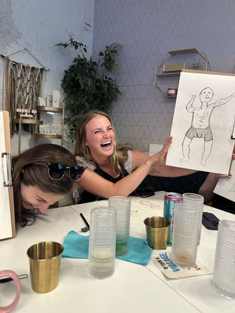 Two friends at an indoor sketching party laughing as one holds up a funny pencil drawing of a muscular man in underwear, with glasses, drink cans and art supplies on the table in a cozy studio setting.