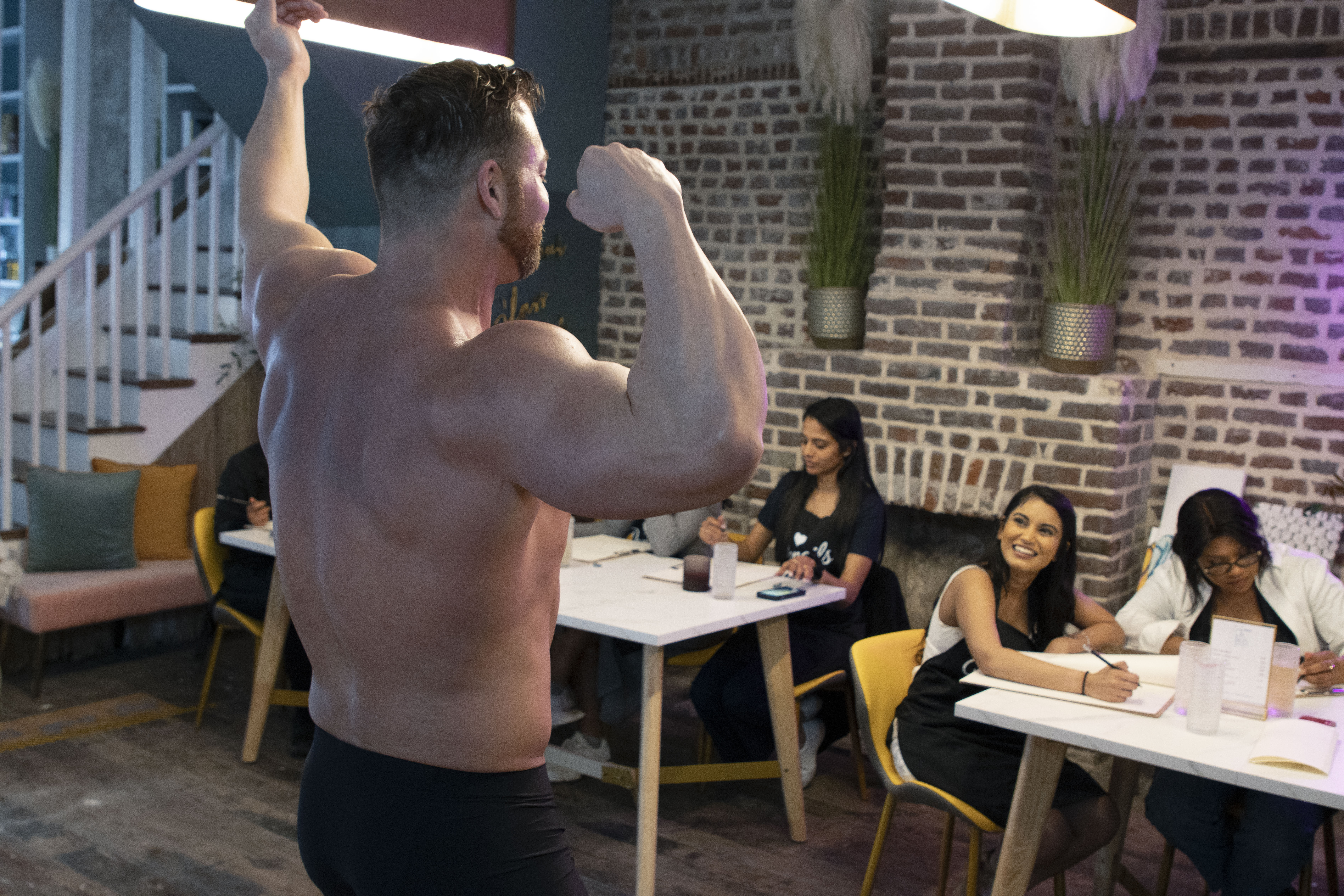 Muscular shirtless male model flexes while seated artists sketch during a figure-drawing workshop in an indoor art studio with exposed brick walls and wooden floors.