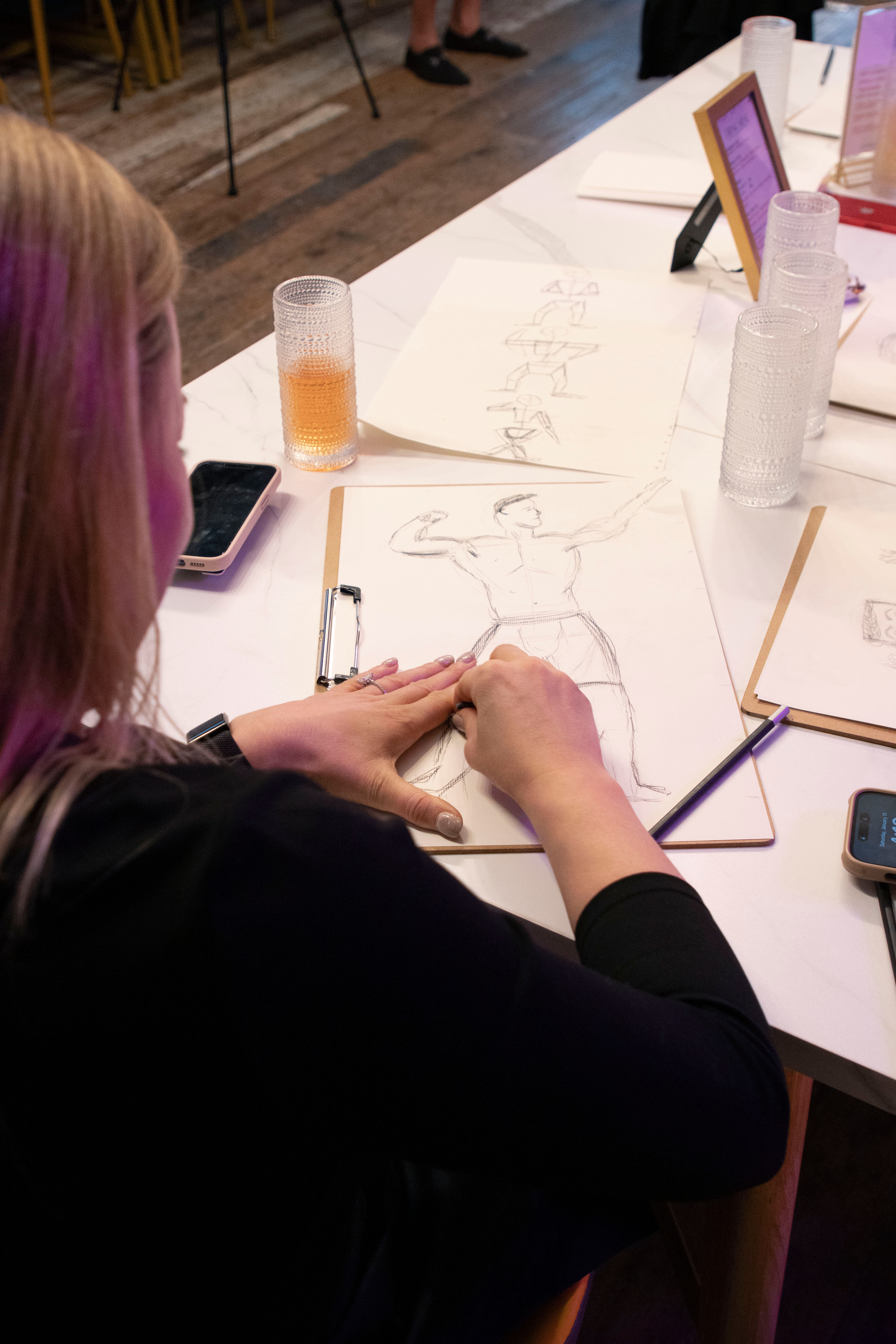 Artist sketching a flexing male figure on a clipboard during a life-drawing workshop, table with pencils, smartphones and textured drinking glasses.