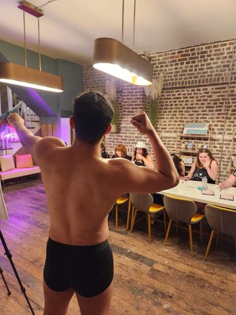 Playful shirtless man in black briefs flexing with his back to the camera in an exposed-brick loft with wooden floors and pendant lights, while a group of women at a long table watch and laugh — indoor party scene.