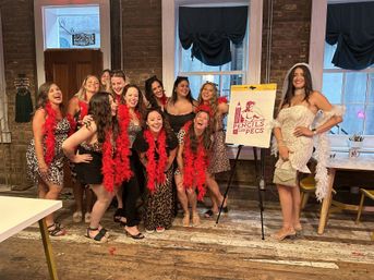 Bachelorette party in a rustic brick-walled art studio — a laughing group of women in leopard-print outfits and red feather boas, the bride in a white lace dress and veil posing beside an easel on worn wooden floors.