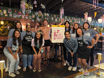 Smiling group of people in matching event shirts and aprons posing around an easel and a shirtless man flexing inside a festive café-bar with hanging disco balls, chandeliers and floral decorations.