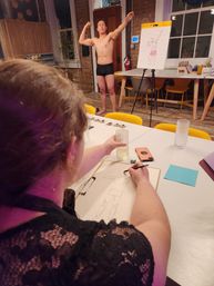 Life drawing class in an urban art studio: a seated artist sketches a shirtless model in black briefs striking a flexing pose, with a clipboard, glass of water and smartphone on the table and an easel, brick wall and yellow chairs in the background.