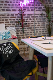 Cozy creative workshop interior with exposed brick wall and pink neon sign, white table set with clipboards, pencils and erasers, yellow chairs draped with black aprons and decorative plants.
