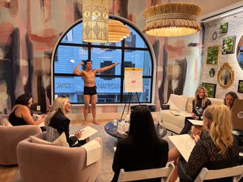 Life-drawing class: women sketching a standing male model in black briefs in a cozy modern studio with an arched window, textured mural wall, and woven pendant lights.