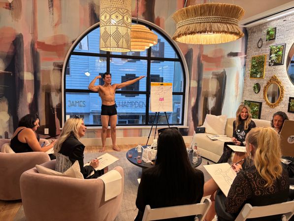 Life-drawing class: women sketching a standing male model in black briefs in a cozy modern studio with an arched window, textured mural wall, and woven pendant lights.