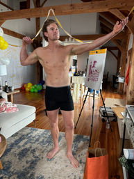 Shirtless man posing in black shorts in a sunlit timber‑beamed loft living room with hardwood floors, balloons and streamers, a couch and rug, and an easel displaying a stylized pencil-and-muscle logo.