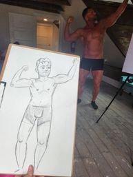 Clipboard showing a quick pencil sketch of a flexing male figure in the foreground, with a shirtless muscular model in black briefs posing in a sunlit loft-style art studio with wooden floor, exposed beams and a tripod easel in the background.
