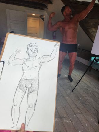 Clipboard showing a quick pencil sketch of a flexing male figure in the foreground, with a shirtless muscular model in black briefs posing in a sunlit loft-style art studio with wooden floor, exposed beams and a tripod easel in the background.