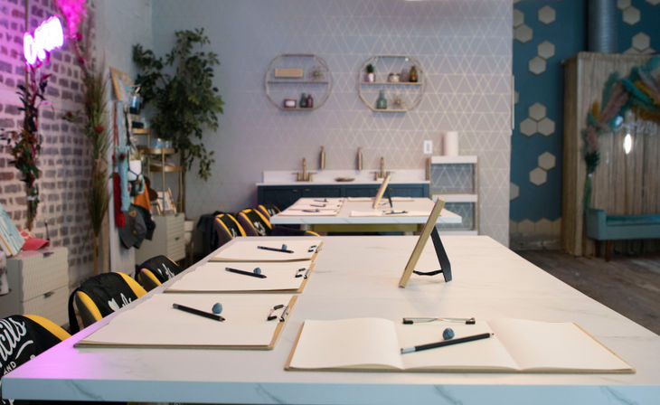 Bright modern art studio workshop with long white tables set for sketching — clipboards, sketchpads, pencils and erasers, black-and-yellow chairs, exposed brick and plant decor.