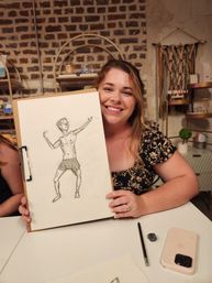 Smiling woman in a floral blouse holding a clipboard with a life-drawing sketch of a male figure at an art studio table, brick wall backdrop with shelves, pencil, eraser and smartphone visible.