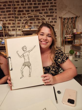 Smiling woman in a floral blouse holding a clipboard with a life-drawing sketch of a male figure at an art studio table, brick wall backdrop with shelves, pencil, eraser and smartphone visible.
