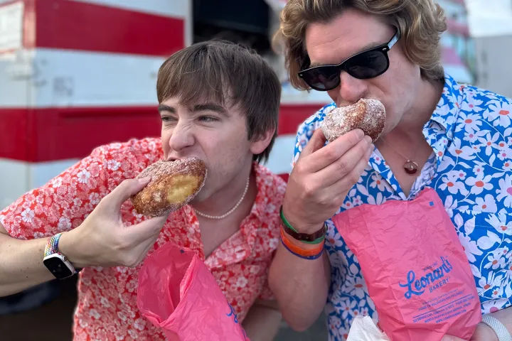 Two people in bright floral shirts enjoying sugar-dusted filled donuts from pink bakery bags in front of a red-striped food truck.
