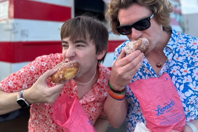Two people in bright floral shirts enjoying sugar-dusted filled donuts from pink bakery bags in front of a red-striped food truck.