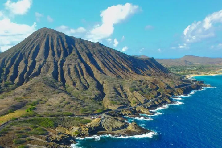 Aerial view of a dramatic volcanic ridge and eroded crater dropping to a turquoise tropical island coastline with rocky cliffs, white surf, and a bright blue sky.