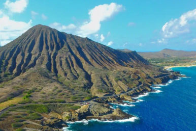 Aerial view of a rugged volcanic coastal ridge with deep radial gullies, rocky cliffs and turquoise ocean waves crashing under a bright blue sky.
