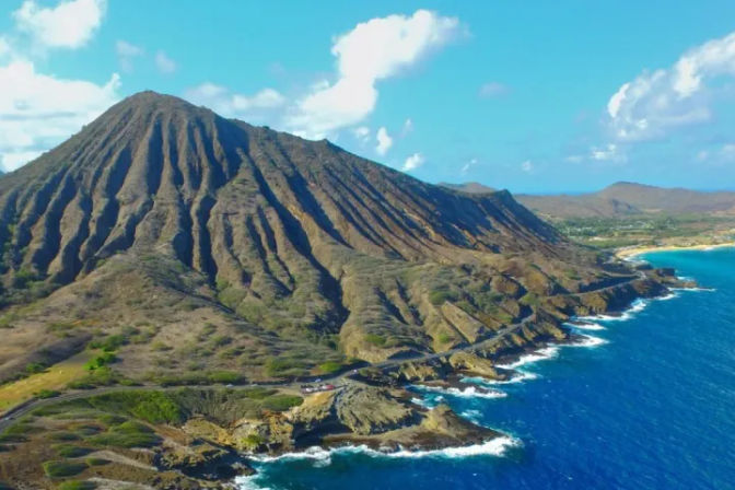 Aerial view of a dramatic volcanic ridge and eroded crater dropping to a turquoise tropical island coastline with rocky cliffs, white surf, and a bright blue sky.