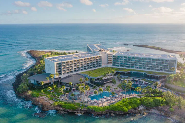 Aerial view of a sunlit oceanfront resort on a rocky tropical peninsula with palm-lined pools, guest balconies, and turquoise sea.