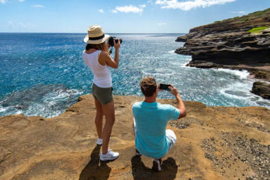 Two travelers on a sunny ocean cliff snapping photos of the bright blue sea and rugged rocky coastline.