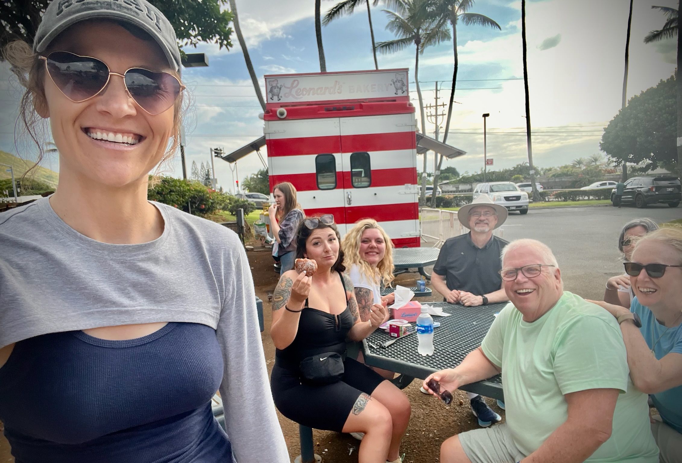 Smiling group of friends and family gathered at an outdoor picnic table by a red-and-white food truck, enjoying donuts and water under palm trees in a sunny tropical parking lot.