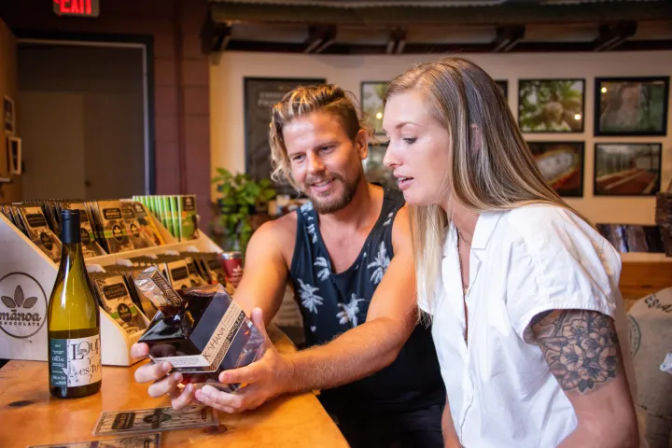 Two shoppers inspect a boxed gourmet chocolate liqueur at a cozy artisan shop counter, with a wine bottle and packaged treats on display