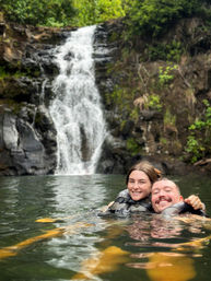 Two smiling people wearing life jackets floating in a natural waterfall pool with rocky cliffs and lush green foliage in the background.
