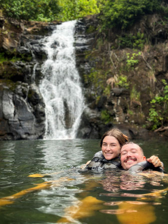 Two smiling people wearing life jackets floating in a natural waterfall pool with rocky cliffs and lush green foliage in the background.