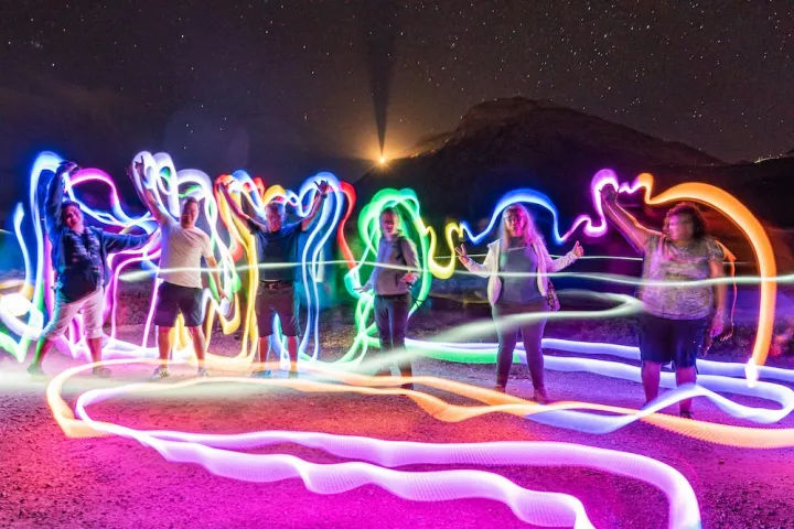 Group of people in a desert at night creating colorful light-painting swirls with LED trails and long-exposure effects beneath a starry sky and silhouetted mountain