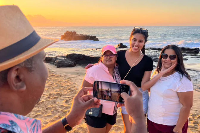 Person in a straw hat photographing three smiling women posing on a sandy beach at golden sunset, ocean waves and rocky outcrops glowing in warm light with the smartphone screen visible.