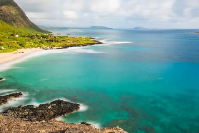 Aerial view of a turquoise coastal bay with a white-sand beach, black rocky shoreline, and lush green cliffs with small islands on the horizon.