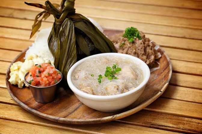 Wooden platter with a leaf-wrapped rice parcel, bowl of clear glass noodle soup topped with scallions, shredded pork, potato salad and fresh pico de gallo