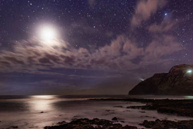 Serene moonlit rocky coastline nightscape with a star-filled sky, wispy clouds, moonlight reflecting on calm ocean and a distant headland with a lighthouse beam.