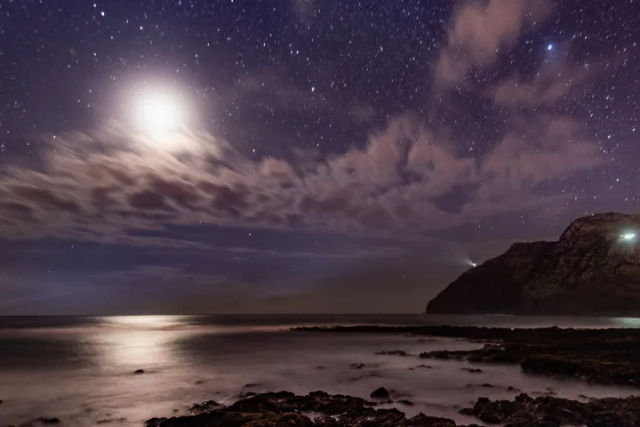 Serene moonlit rocky coastline nightscape with a star-filled sky, wispy clouds, moonlight reflecting on calm ocean and a distant headland with a lighthouse beam.