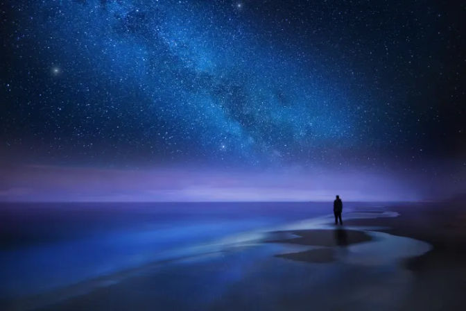 Lone silhouette standing on a tranquil beach at night beneath a vivid blue Milky Way and star-filled sky, reflective shoreline and gentle waves