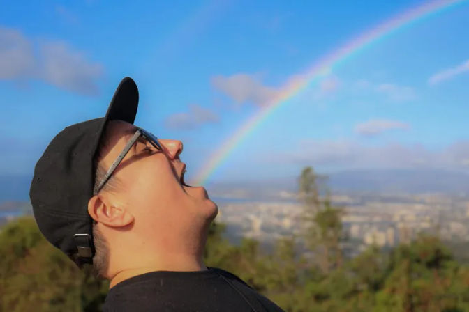 Person wearing a black cap and glasses on a hilltop pretending to bite a bright rainbow arching over a city skyline under a blue sky.