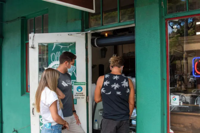 Three people pausing at a teal-green storefront entrance in casual summer clothes (jeans, t-shirt, palm-print tank), one wearing a face mask, next to a sign asking customers to wear masks.
