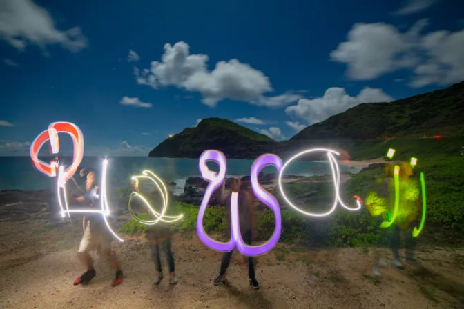 Long-exposure light painting on a tropical coastal cove at night — playful neon scribbles and blurred figures with silhouetted hills and a cloudy sky.
