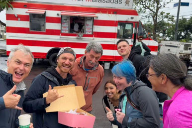 Smiling group of people posing with an open pink box of pastries, giving thumbs-up and shaka signs in front of a red-and-white striped food truck on a wet street.