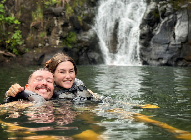 Two smiling people wearing life jackets floating in a natural pool beneath a small waterfall, surrounded by rocky cliffs and green foliage.