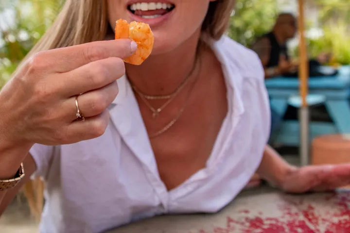 Smiling person in a white shirt holding a cooked shrimp near their mouth at an outdoor patio picnic table — casual seafood dining with summer vibes