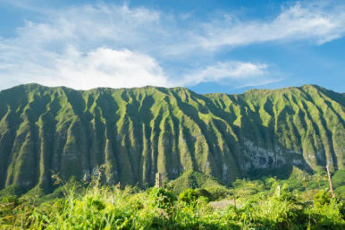 Dramatic lush green tropical mountain ridges with deep vertical grooves under a bright blue sky, wild grasses and valley in the foreground.