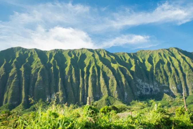 Dramatic lush green tropical mountain ridges with deep vertical grooves under a bright blue sky, wild grasses and valley in the foreground.
