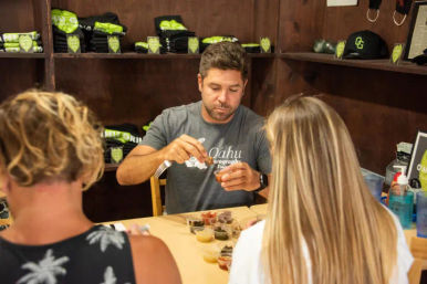 Man scooping small sauce samples for two customers at an indoor tasting table with island-themed merchandise on shelves — Oahu tasting.
